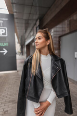 Trendy urban female port of beautiful young woman model in leather black rock jacket with white dress walks in the city