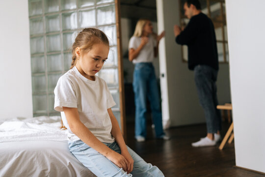 Portrait of depressed cute little daughter sadness looking down sitting on couch during parents quarrelling and fighting in living room on background. Concept of family problems, conflict, crisis.
