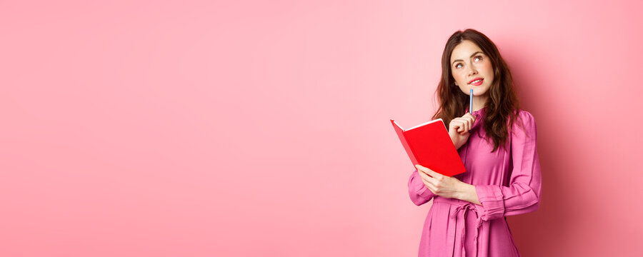 Beautiful young woman look thoughtful, writing in notebook, holding planner or diary, plan her schedule, standing against pink background