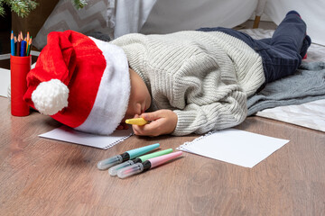 A child fell asleep while writing a wish list for Santa. A little boy in a Santa hat sleeps on the...