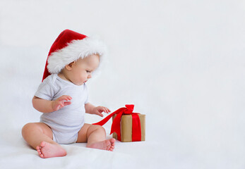 A little boy reaches for a gift. A beautiful child in a Santa Claus hat sits on a white background. Copy space.