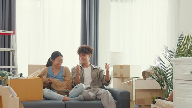 Young happy Asian couple sit on sofa couch use design thinking pointing wall get idea decoration room in living room at home. Husband wife move new house, Apartment Rent And Ownership Concept.