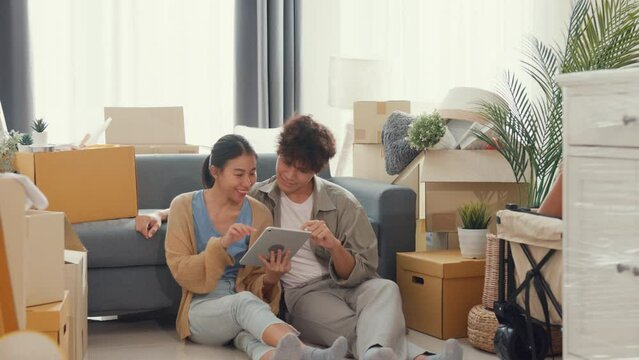 Young happy Asia couple sit on sofa couch use digital tablet online shopping furniture from decoration store in living room at home. Husband wife move new house, Apartment Rent And Ownership Concept.