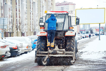 Tractor with brush and scraper removes snow at city street, clears sidewalk during heavy snowfall at winter day. Tractor clearing snowy streets with snowplough and rotary brush. Vehicle sweeps snow