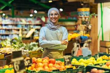 Portrait woman in a hijab as a seller in a supermarket store, a saleswoman with arms crossed smiles and looks at the camera, an apron sells apples and fruits, a Muslim woman is satisfied in glasses.