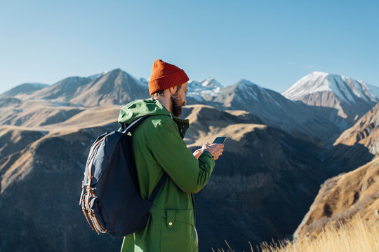 a man with a backpack in a green raincoat stands and looks at the phone in the mountains