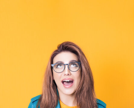 Surprised young woman looking up to free space - copy space for your product or text, wearing blue leather jacket, isolated on yellow background
