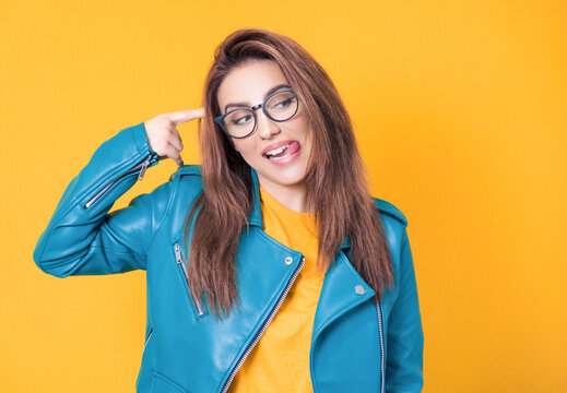 Funny woman makes finger gun gesture in temple, pretends committing suicide, wearing blue leather jacket, isolated on yellow background. Silly girl sticking out tongue