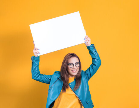 Beautiful women with surprised face holding white speech bubble, wearing blue leather jacket, isolated on yellow background. Female person shows blank empty paper billboard with blank space