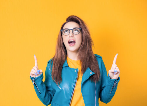 Young woman looking up and pointing to shows your space for product, promoter girl, wearing blue leather jacket, isolated on yellow background