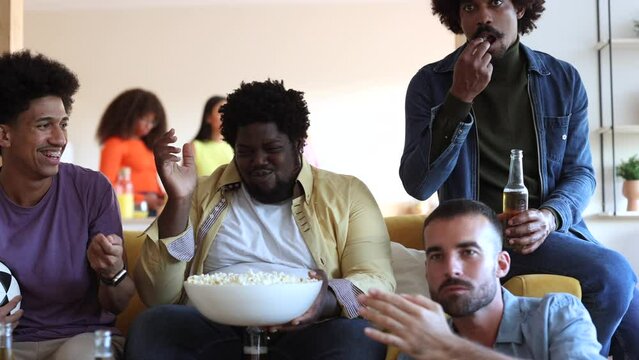 Multiracial Group Of Friends Eating Popcorn And Drinking Beers Sitting On The Sofa Concentrating While Watching A Soccer Game On TV. Focus On Overweight Plus Size American African Man