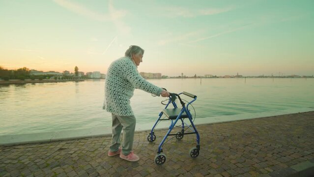 Grandmother using rollator with short grey hair wearing warm woolen cardigan strolls on cobbled road along Venice lagoon in Sottomarina