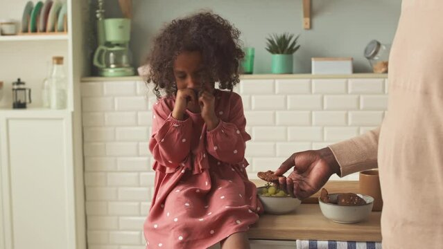 African American little girl in cute punk polka dot dress sitting on wooden kitchen table and crying while her father trying to calm her down and offering some food
