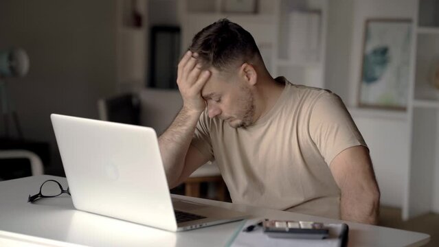 Male hipster freelance businessman tired working at laptop computer, has pain in head taking off glasses and touching eyes. Exhausted working at screen has eye strain, dry eyes and need eye drops
