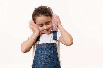 Fashionable little girl in blue denim overalls, holding her hands on furry plush earmuffs, enjoying her new stylish winter accessory. Protect your ears from the cold, isolated over white background