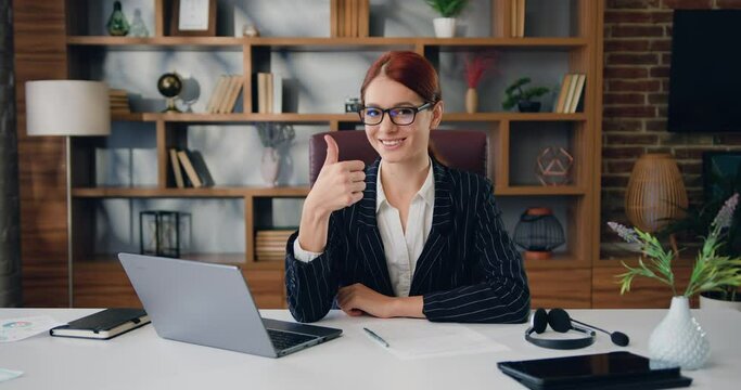 Close-up portrait of smiling caucasian businesswoman in glasses with red hair showing thumb up, recommending online computer program while sitting in front of laptop at workplace in modern office