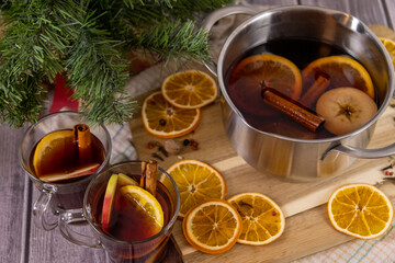 Mulled wine in glass mugs placed on wooden table with hot pan with red wine, cinnamon sticks, spice and orange fruit, Christmas decorations