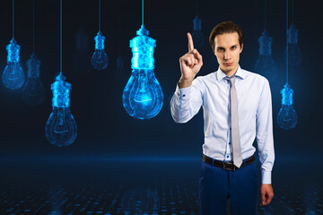 Attractive young european businessman pointing at glowing digital lamp hologram on dark background. Idea, innovation and future concept.