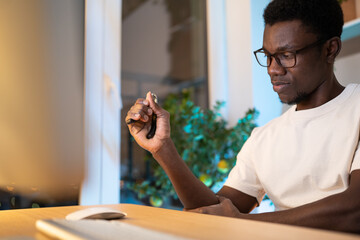 Fototapeta na wymiar Young black worker in glasses trains hand muscles sitting at desk in living room at home. African American man in white t-shirt takes care of wrist using expander during long time work on computer