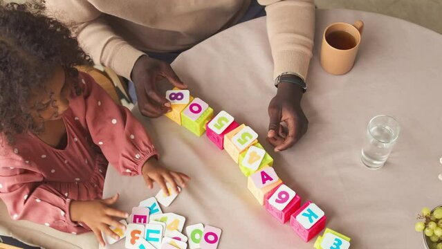 From above shot of African American little girl together with dad learning alphabet together at table using colorful magnetic letters and numbers