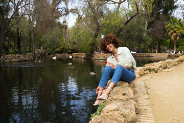 Beautiful woman with curly hair sitting on a duck and swan pond in the park. The woman is happy and holds a red flower in her hand as she looks at it excitedly.