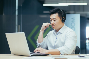 Fototapeta premium Portrait of young Asian male consultant in support service. Sitting in the office at the table, wearing headphones and a microphone, talking with clients online from a laptop.