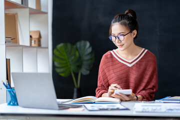 Portrait of an Asian businesswoman pressing a calculator, looking at a notebook, analyzing data, and reports, preparing presentations, marketing, and finance for sales in online shopping systems.