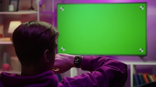 Man using smartwatch, watching television with chroma key close-up. Young guy sitting on sofa and wearing fitness bracelet, checking pulse. 