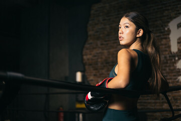 Young Asian woman leaning on the ropes of a boxing ring while resting, youth boxing.