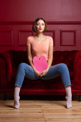 Women in jeans with open legs sitting straight back on couch and holding heart shapes