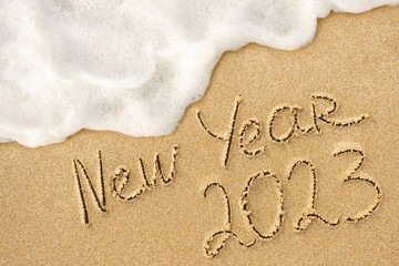 New Year 2023 written words on the sand, beach and foamy ocean wave on background. Winter holidays...