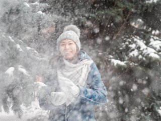 Smiling woman in cable knitted hat is playing with snow. Fun in park between snowy fir trees. Woman laughs as she is throwing snowball. Cold season.