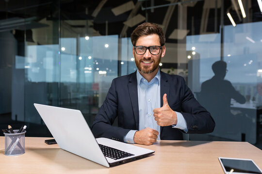Portrait of mature successful investor, businessman in business suit working inside office using laptop, senior male boss looking at camera smiling and showing thumbs up.