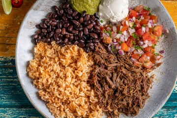 Top-view of a barbacoa burrito bowl, meat rice black beans sour cream guacamole chopped vegetables