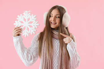 Young woman in warm ear muffs pointing at big snowflake on pink background