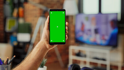 Male freelancer holding smartphone with green screen display in living room, using isolated template with chroma key and mockup. Analyzing blank copyspace background on mobile phone.