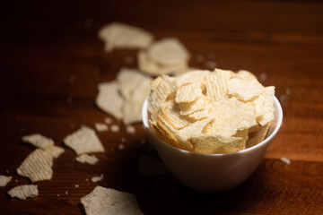 white bowl with potato ribbed ruddy appetizing chips on a blurred background of a dark wooden table with scattered chips