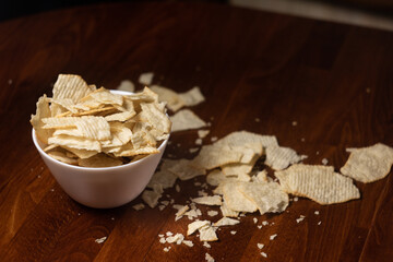 scattered chips and a white bowl filled with potato ribbed chips on a dark wooden table background
