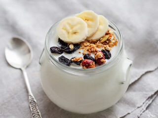 Natural homemade yogurt in glass jar. Healthy food for breakfast with muesli. Jar with granola and banana slices on linen tablecloth on linen napkin.