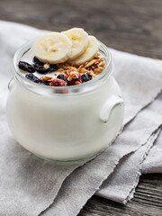 Natural homemade yogurt in a glass jar. Healthy food for breakfast with muesli. Jar with granola and banana slices on linen tablecloth on wooden table.