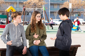 Group of three cheerful children talking and laughing on bench on playground