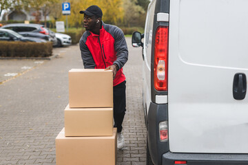 Young adult black delivery man standing next to white van with unloaded parcels stacked on trolley....
