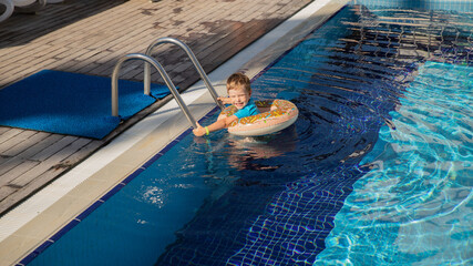 A little happy boy bathes in a doughnut lifebuoy in clear water in swimming pool in sunny weather