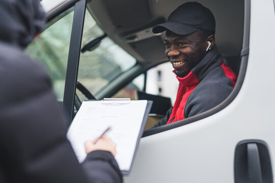 Happy positive Black courier joking around with unrecognizable person taking a delivery. Unrecognizable person signing delivery documents standing next to white delivery truck. High quality photo