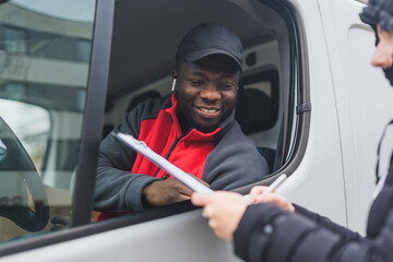 Person signing papers held out window of white van by young adult black delivery man wearing red...