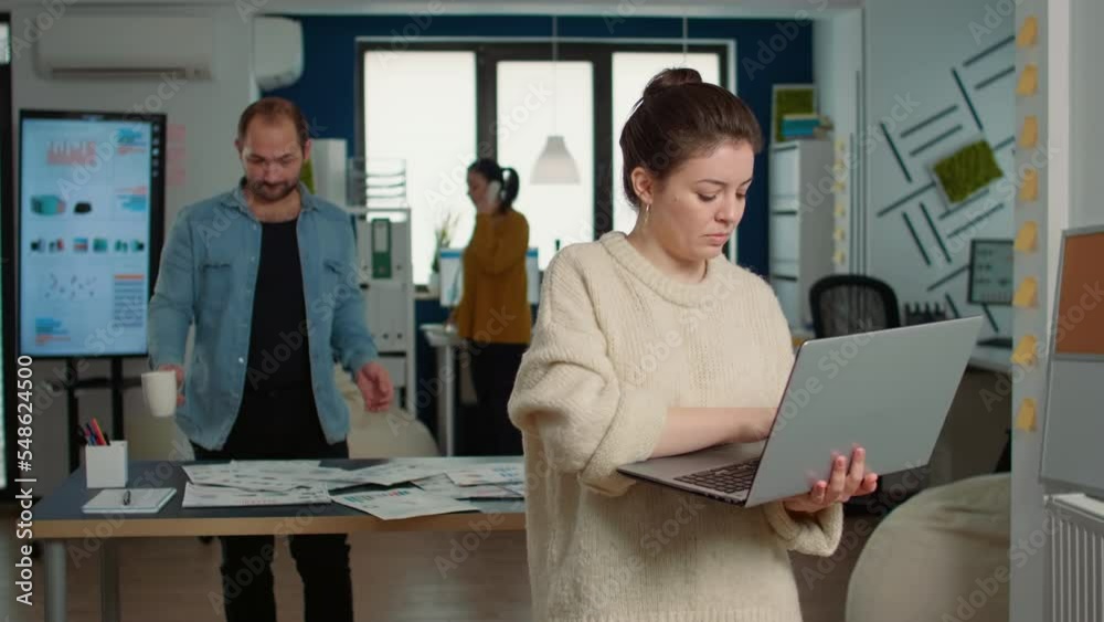 Wall mural Portrait of casual woman using laptop and smiling at camera while coworker holding cup looks at papers with charts. Confident startup employee working relaxed in busy office of small company.