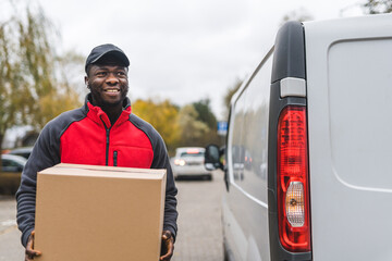 Black young adult delivery man wearing work uniform smiling carrying cardboard box parcel unloaded...