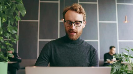 A young red-haired guy, a programmer or an entrepreneur in glasses, in a stylish cafe Looks at the camera and smiles. Freelancer works remotely. Online communication. small business.