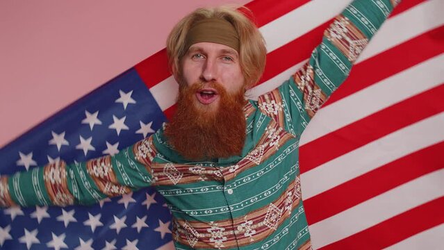 Hippie redhead bearded man in pattern shirt waving and wrapping in American USA flag, celebrating, human rights, freedoms. Independence day. Hipster ginger guy boy isolated on pink studio background