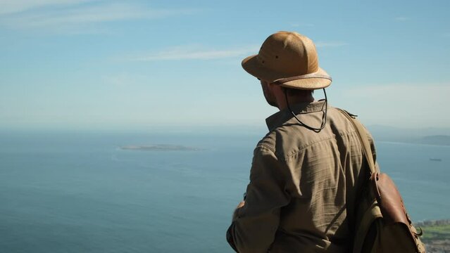 a male traveler in a sun hat points a finger at a famous island in the ocean. hiker man stands on a high mountain against the backdrop of the atlantic ocean. Robben Island, Cape Town, South Africa.